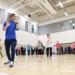 A man wearing a blue t-shirt is leading a tai-chi class with a group of older people.