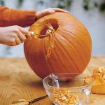 A picture of a pumpkin with the top cut off it and someone scooping out the seeds from inside.