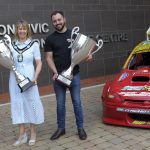 Male and female holding trophies beside a racing car.