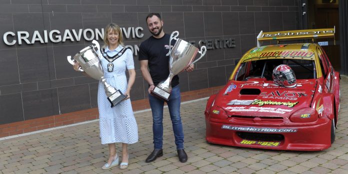 Male and female holding trophies beside a racing car.
