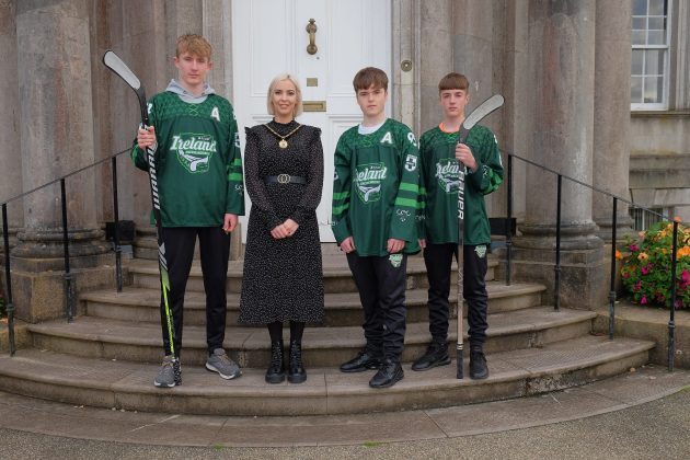 Three males in hockey clothing and holding two hockey stick standing with a female council official.