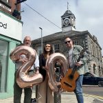 Three individuals standing in front of a stone building with a clock tower, holding balloons shaped as the numbers 20. One person also holds a guitar.