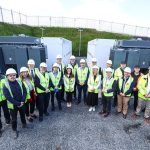Group of people in high vis vests and whit hard hats standing at a work site.