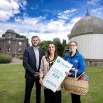 Three people standing outside an observatory with a large telescope dome. One individual holds a sign and another holds a picnic basket. The background features lush greenery and the observatory building.