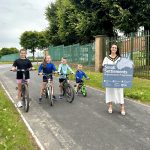 Four children on bicycles smile as they ride along a paved road next to a grassy area, accompanied by an adult holding a sign reading "Small Settlements Regeneration Programme." Trees and a green fence line the background.