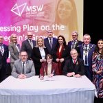 A group of dignitaries standing with a table in front and signs for Mid South West on a screen behind them.