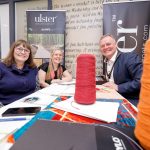 Three individuals sitting at a table with large colorful spools of thread, smiling at the camera, in front of an "Ulster Carpets" banner.