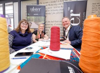 Three individuals sitting at a table with large colorful spools of thread, smiling at the camera, in front of an "Ulster Carpets" banner.
