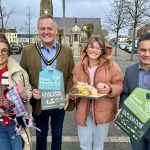 Four individuals holding promotional items for "Food Heartland" standing in Lurgan town square with buildings in the background. Two individuals are holding products - buns, and pet accessories.