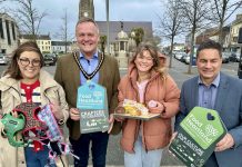 Four individuals holding promotional items for "Food Heartland" standing in Lurgan town square with buildings in the background. Two individuals are holding products - buns, and pet accessories.