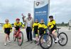 Five young triathletes are pictured with the lord mayor beside South Lake in Craigavon. Two children are on bicycles and the lord mayor is holding a megaphone, raised in the air.