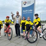 Five young triathletes are pictured with the lord mayor beside South Lake in Craigavon. Two children are on bicycles and the lord mayor is holding a megaphone, raised in the air.
