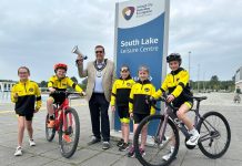 Five young triathletes are pictured with the lord mayor beside South Lake in Craigavon. Two children are on bicycles and the lord mayor is holding a megaphone, raised in the air.
