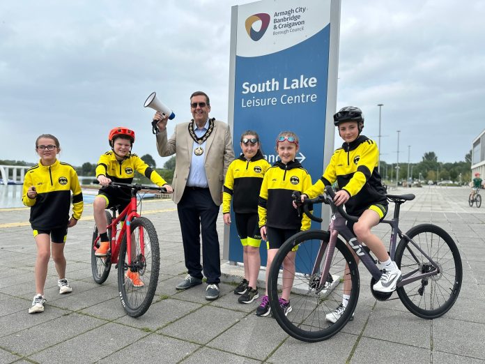 SL Jr Triathlon 2025 Five young triathletes are pictured with the lord mayor beside South Lake in Craigavon. Two children are on bicycles and the lord mayor is holding a megaphone, raised in the air.