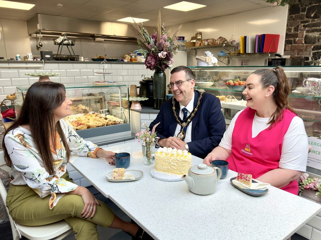 Three people sitting around a table in a cafe, chatting and laughing. There are cakes and a teapot on the table.