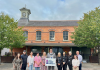A group of people standing in Dromore Market Square with the hall and clock in the background. In the middle of the group, the Lord Mayor is holding a sign saying High Street Heroes NI 2025.