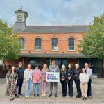A group of people standing in Dromore Market Square with the hall and clock in the background. In the middle of the group, the Lord Mayor is holding a sign saying High Street Heroes NI 2025.