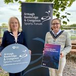 Two women standing in front of an ABC Sports Forum pop up banner and holding signs to promote the Coach Education and Club Development programme.
