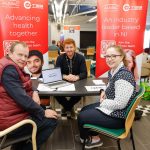 Three people sitting at a job fair stand for Almac in Portadown, surrounded by branded signs.