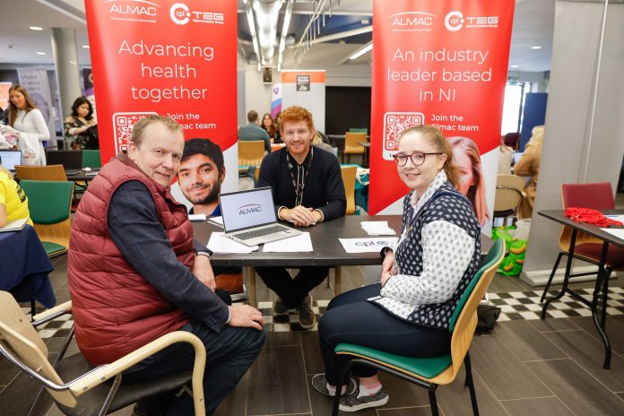 0049_ABC_LMP_PortadownJob Three people sitting at a job fair stand for Almac in Portadown, surrounded by branded signs.
