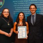 Gymnastics continues to RISE at Dromore Community Centre Three people standing in front of a large screen at an awards ceremony. the middle person is holding a certificate.