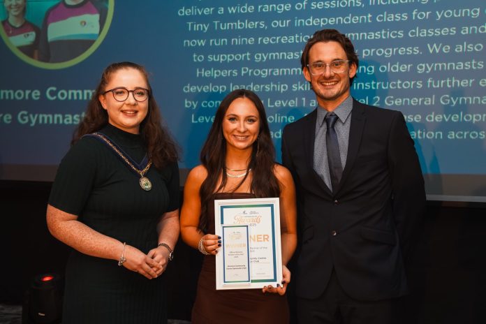 NI- Awards_0152 Three people standing in front of a large screen at an awards ceremony. the middle person is holding a certificate.