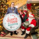 Lord Mayor standing in front of a Christmas tree with Santa Claus holding a present, seated, and an elf in between holding candy canes. The Lord Mayor is holding a circular sign saying Santa on Tour across Armagh City, Banbridge and Craigavon.