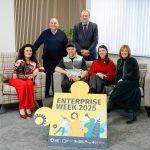 A group of people, some sitting on a sofa and some standing behind. There is a sign in front of them which reads, Enterprise Week 2025.
