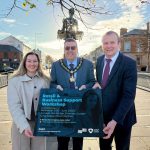 Three people standing on Lurgan Plaza holding a sign that says Retail & Business Support Workshops