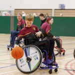 A boy is a wheelchair bouncing a basketball in a sports hall