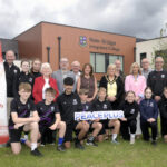 Local school-club partnership to deliver PEACEPLUS funded sports floodlighting project in Loughbrickland Large group of people standing outside New-Bridge Integrated College with some students kneeling in the front row and holding a PEACEPLUS sign.