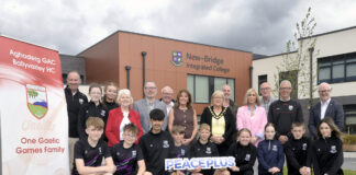 Local school-club partnership to deliver PEACEPLUS funded sports floodlighting project in Loughbrickland Large group of people standing outside New-Bridge Integrated College with some students kneeling in the front row and holding a PEACEPLUS sign.