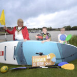 the lord mayor and 2 children standing beside a lake with a paddleboard and sports raquets.
