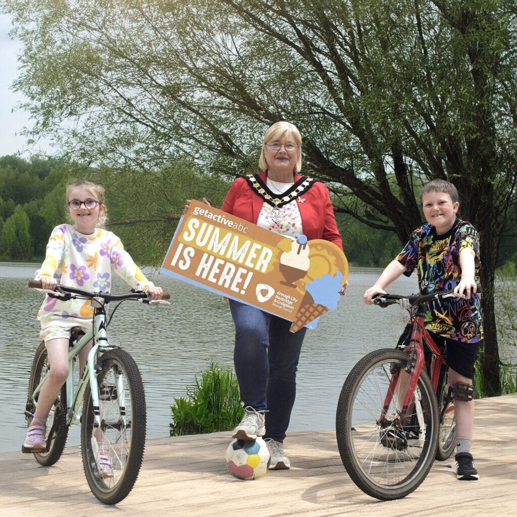The lord mayor is holding a 'summer is here' sign with children on bicycles at either side
