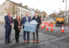 Four people standing on a street with construction works to their left, holding a sign saying Small Settlements.